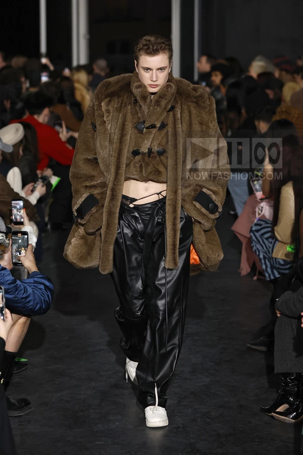Model walking a runway wearing a cropped brown faux-fur coat with toggle closures and black leather trousers.