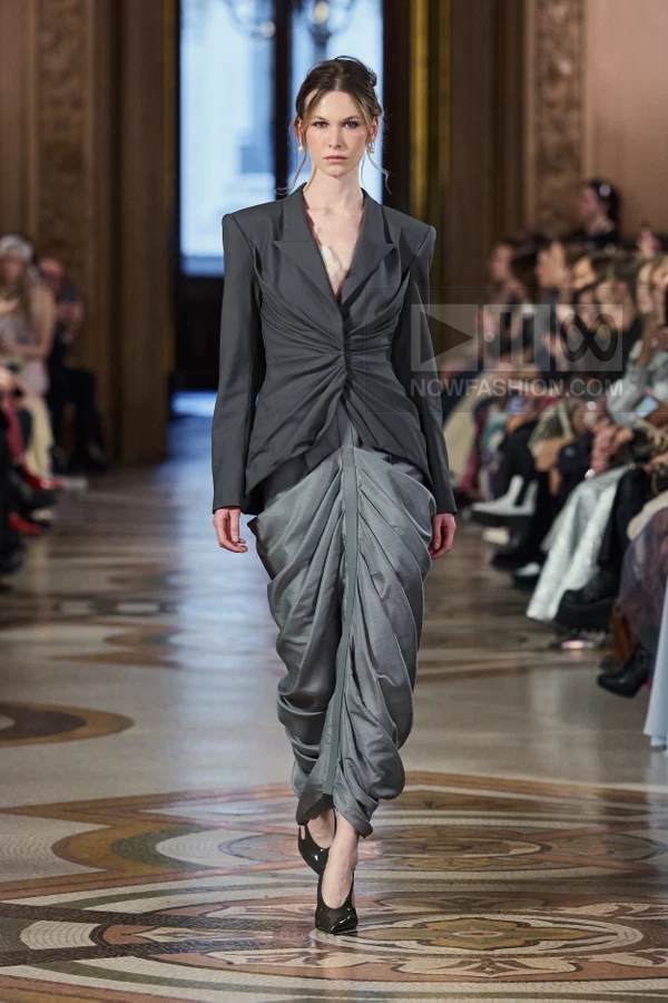 Model walking in a charcoal gray blazer with center-front twist and a matching draped, voluminous skirt on a runway.