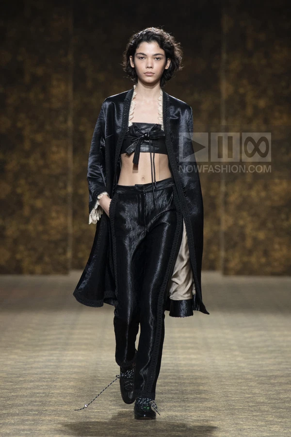 Model on runway wearing a black satin coat over a black leather bandeau top and matching trousers with lace trim.