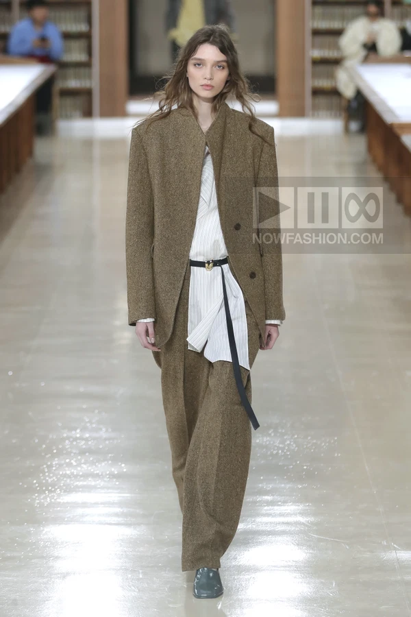 Model wearing a brown tweed suit with a white pinstriped shirt and a thin black belt on a library runway.