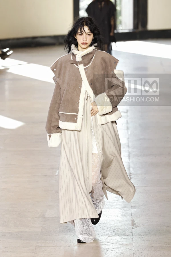 Model walking in a brown shearling-lined jacket over a long pinstriped coat and white lace trousers on a runway.