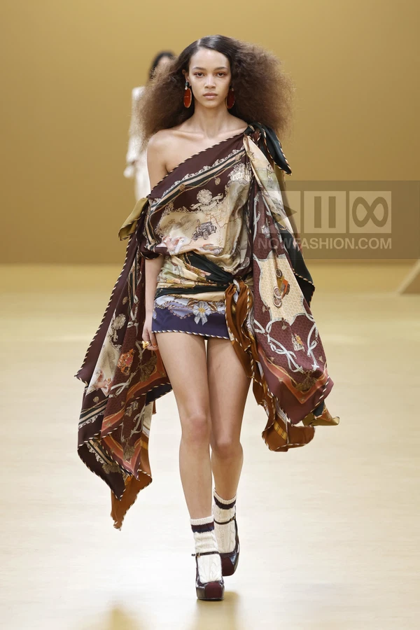 Model walking in a brown printed silk scarf-style mini dress with one shoulder, paired with white socks and dark heels.